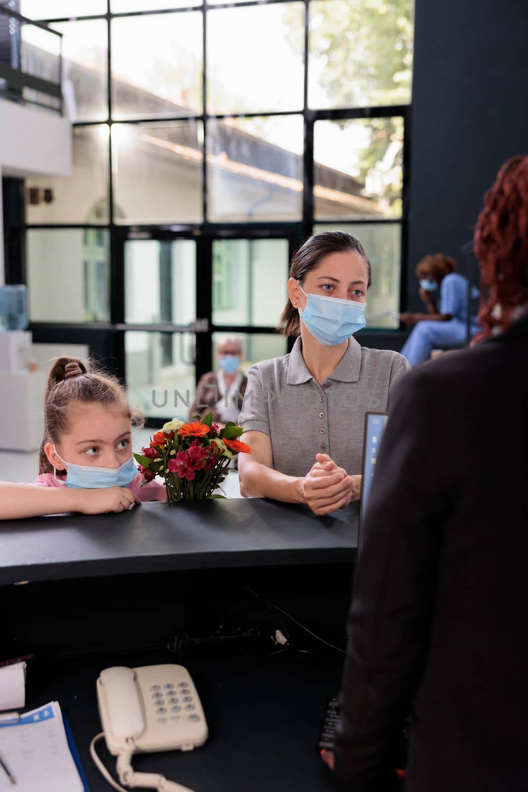 Mother and kid wearing protection face mask against covid19 while discussing medical insurance with receptionist during checkup visit examination in hospital reception. Medicine service and concept