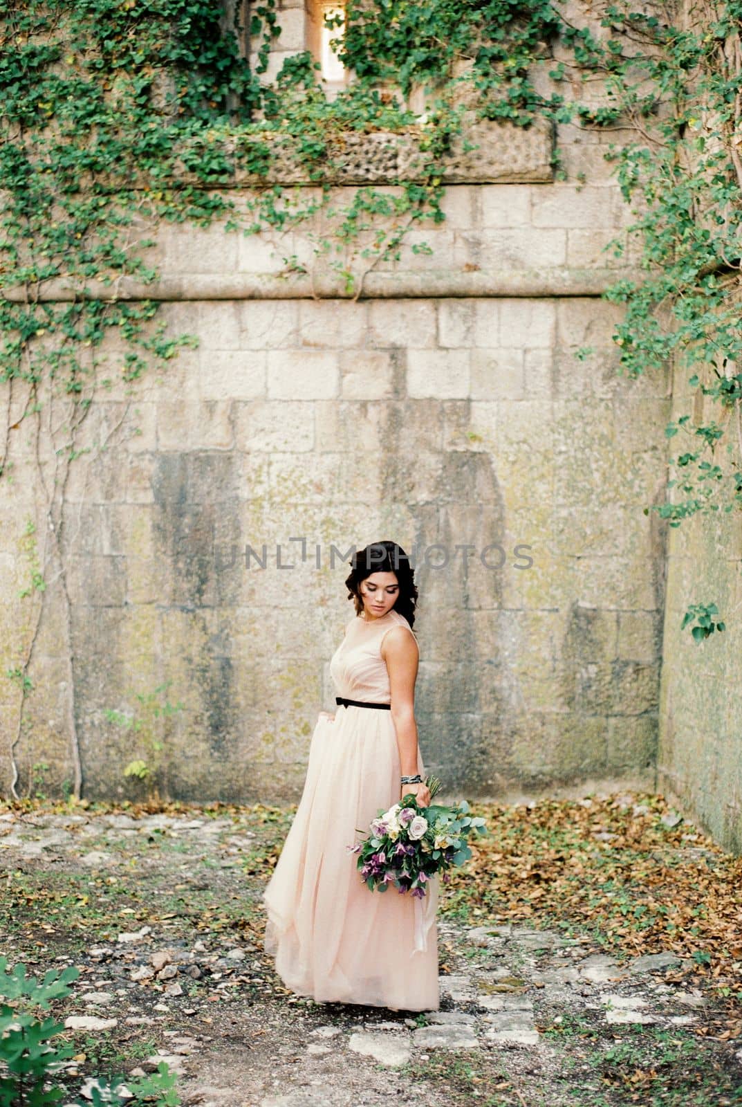 Bride in a dress stands near a stone wall and looks at a bouquet in her hand. High quality photo