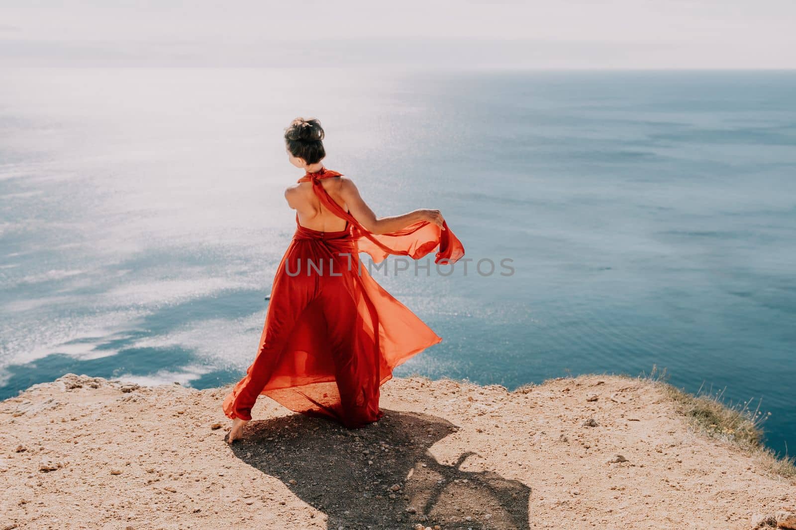 Woman in red dress on sea. Side view a Young beautiful sensual woman in a red long dress posing on a rock high above the sea on sunset. Girl on the nature on blue sky background. Fashion photo. by panophotograph