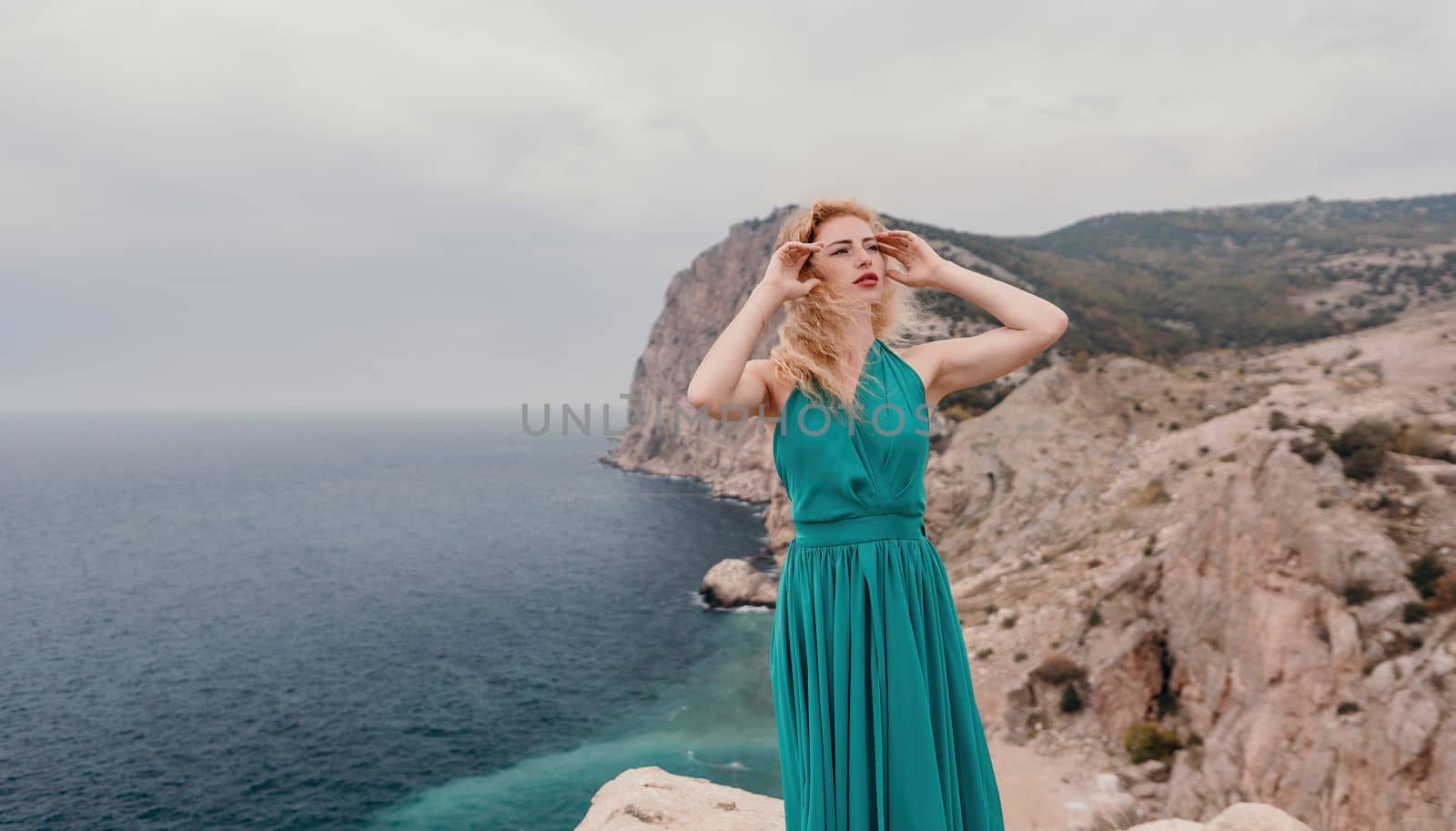 Side view a Young beautiful sensual woman in a mint long dress posing on a volcanic rock high above the sea during sunset. Girl on the nature on overcast sky background. Fashion photo