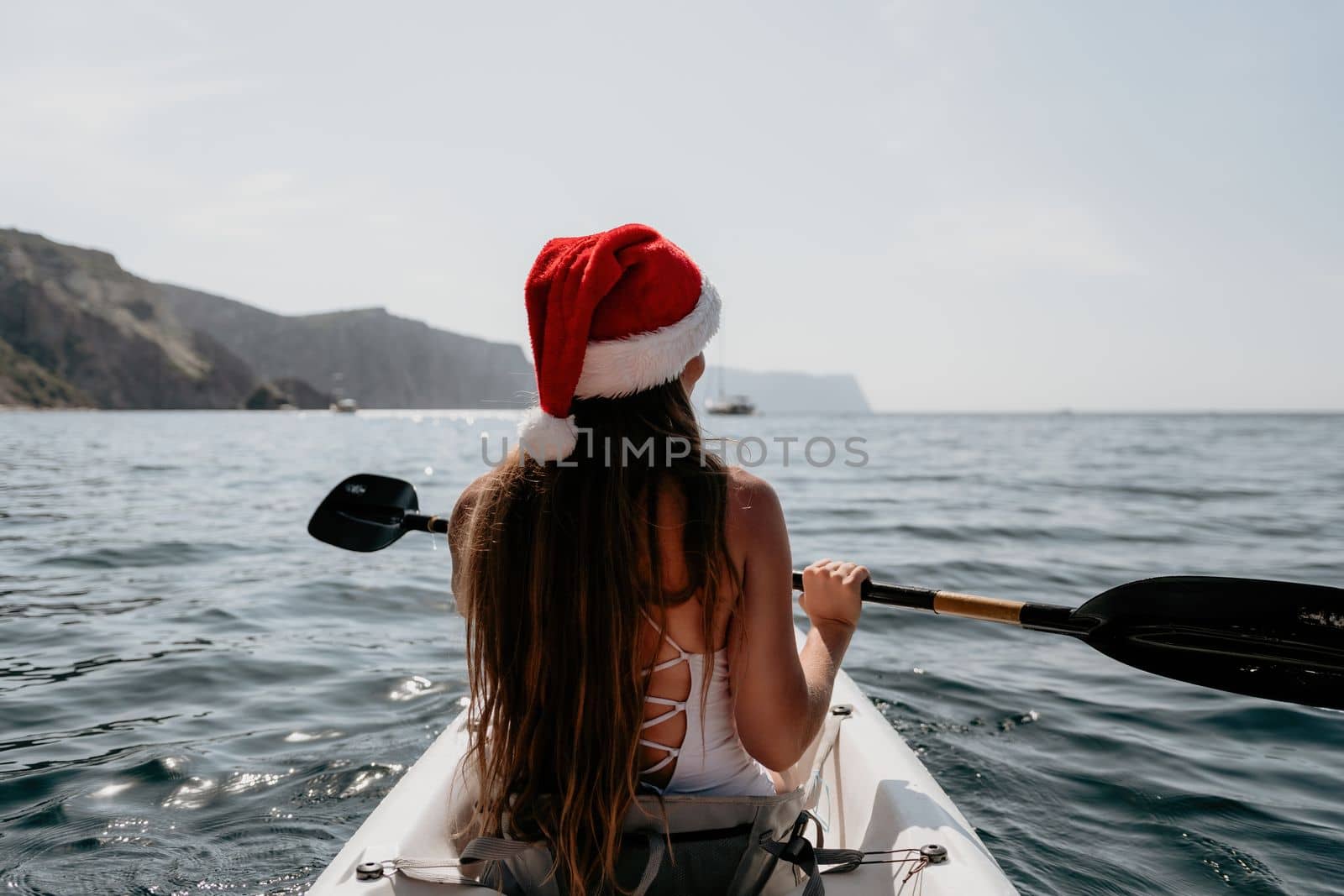 Woman in kayak back view. Happy young woman in Santa hat floating in kayak on calm sea. Summer holiday vacation and cheerful female people relaxing having fun on the boat. by panophotograph