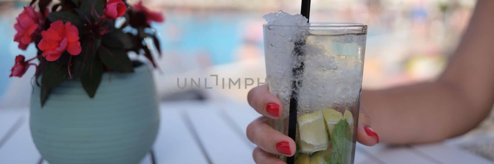 Beautiful glass with refreshing cocktail in woman hand on background of pool. Summer cold drinks concept