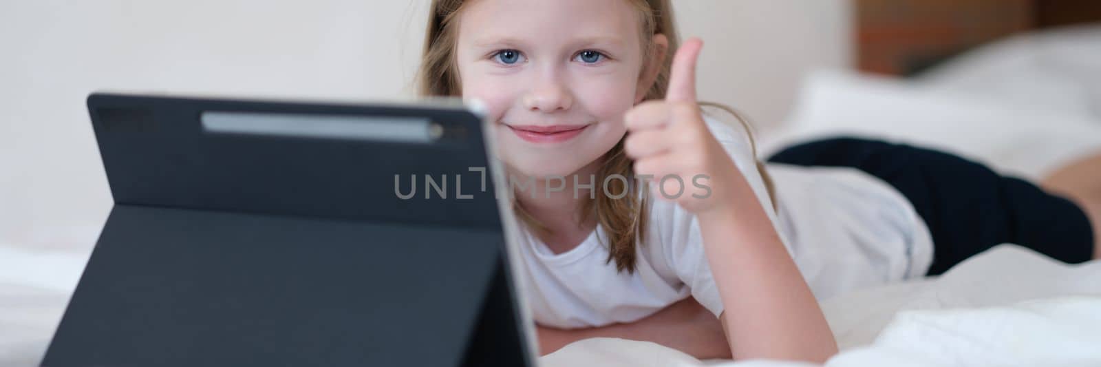 Cheerful child girl showing thumb up lying on bed with tablet. Children rest at home with modern gadgets