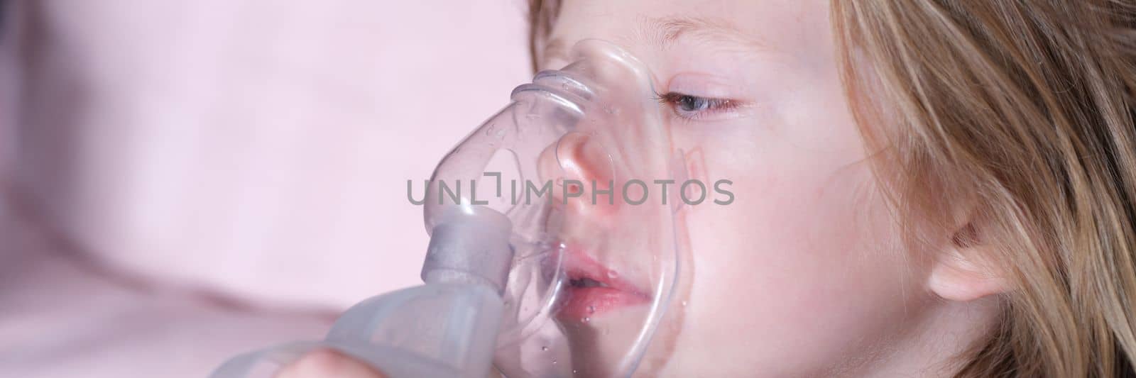 Sick girl in oxygen mask lies on hospital bed. Closeup portrait of child breathing in oxygen mask and lying in bed in pediatric ward