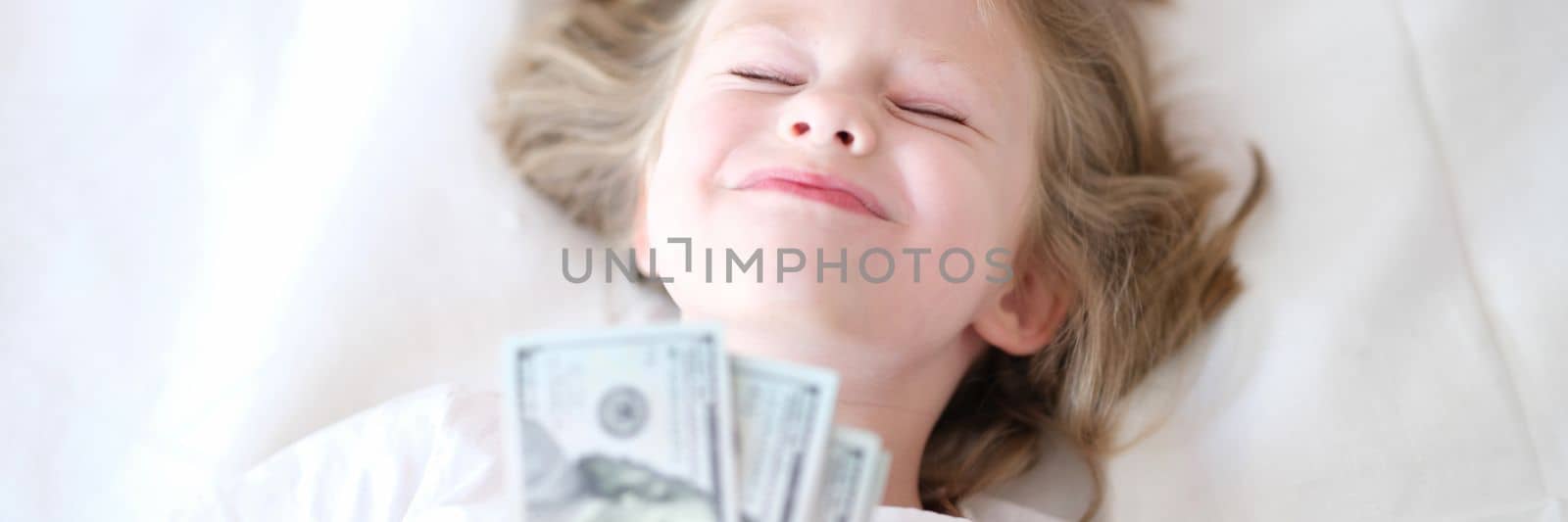 Happy girl holding cash dollars lies on bed with eyes closed. Childish dreams and lot of money