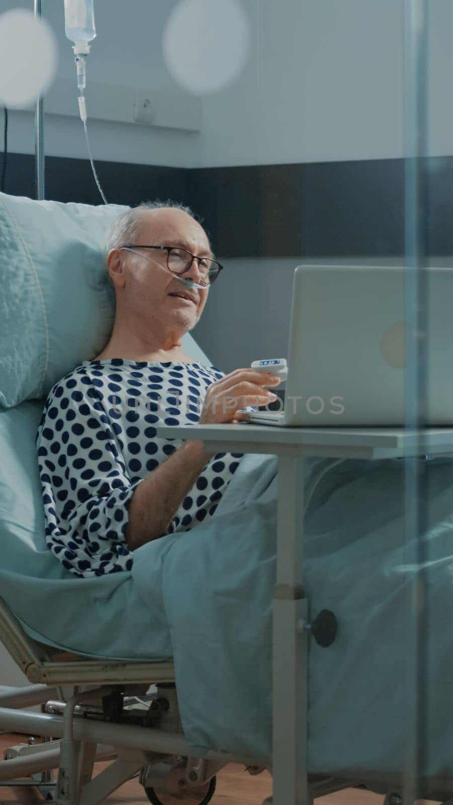Patient in hospital ward talking on videocall with family using laptop. Sick old man with breathing problems wears nasal oxygen tube and oximeter while operating on modern computer