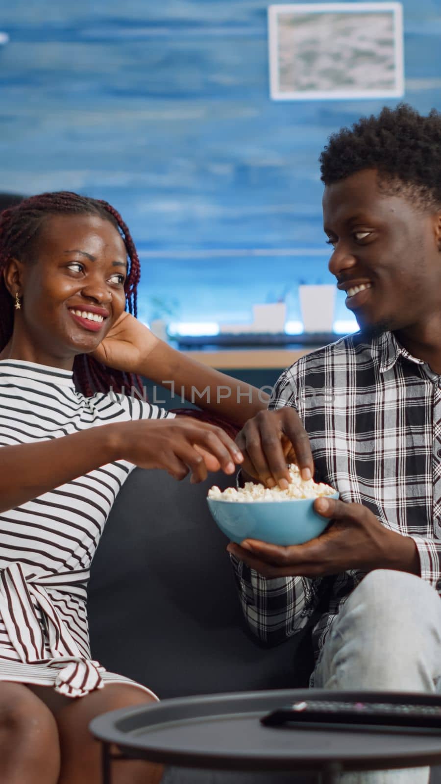 POV of black people having fun while watching TV in living room. African american couple laughing at comedy looking at camera. Cheerful lovers of african ethnicity eating popcorn