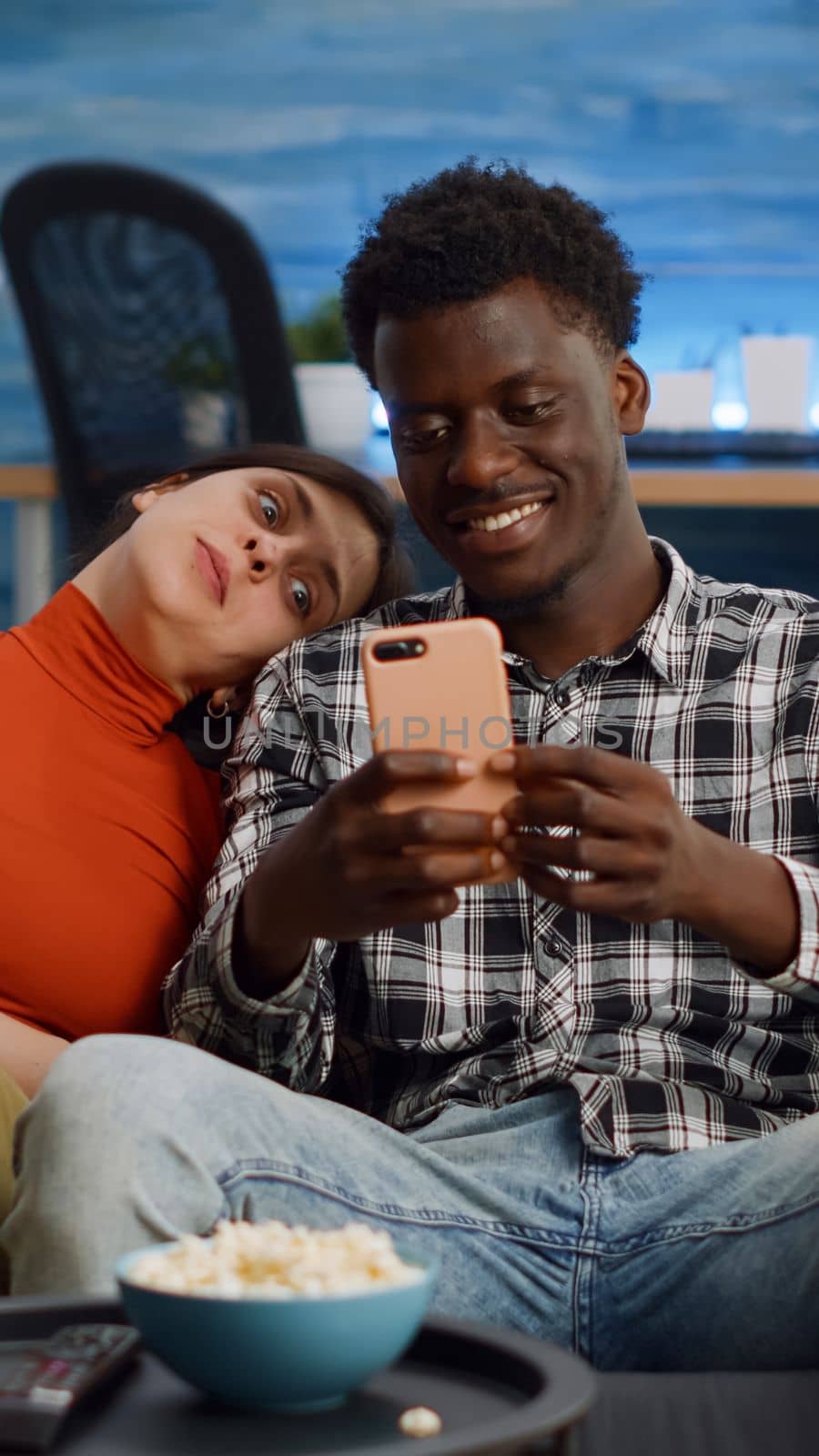 Joyful interracial couple taking selfies with smartphone while sitting together on living room sofa. Cheerful multi ethnic people having fun with pictures and technology at home