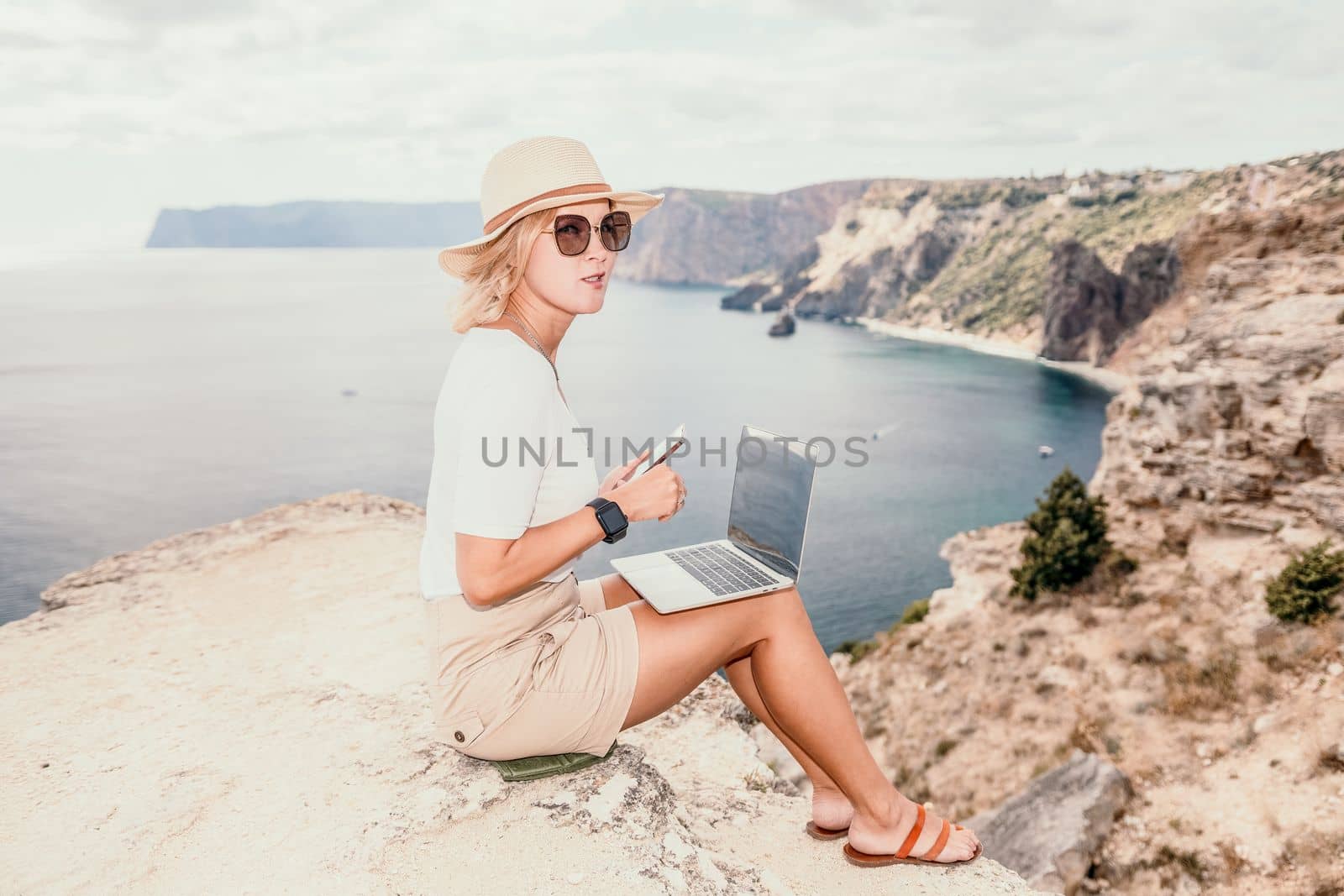 Digital nomad, Business woman working on laptop by the sea. Pretty lady typing on computer by the sea at sunset, makes a business transaction online from a distance. Freelance, remote work on vacation by panophotograph