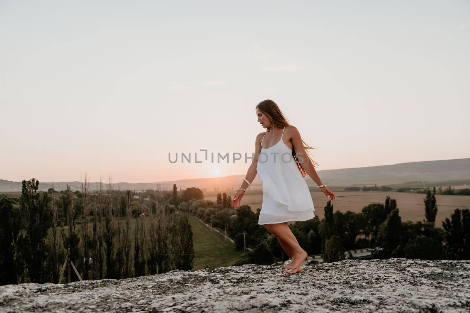 Happy woman standing with her back on the sunset in nature in summer with open hands. Romantic beautiful bride in white boho dress posing with mountains on sunset by panophotograph