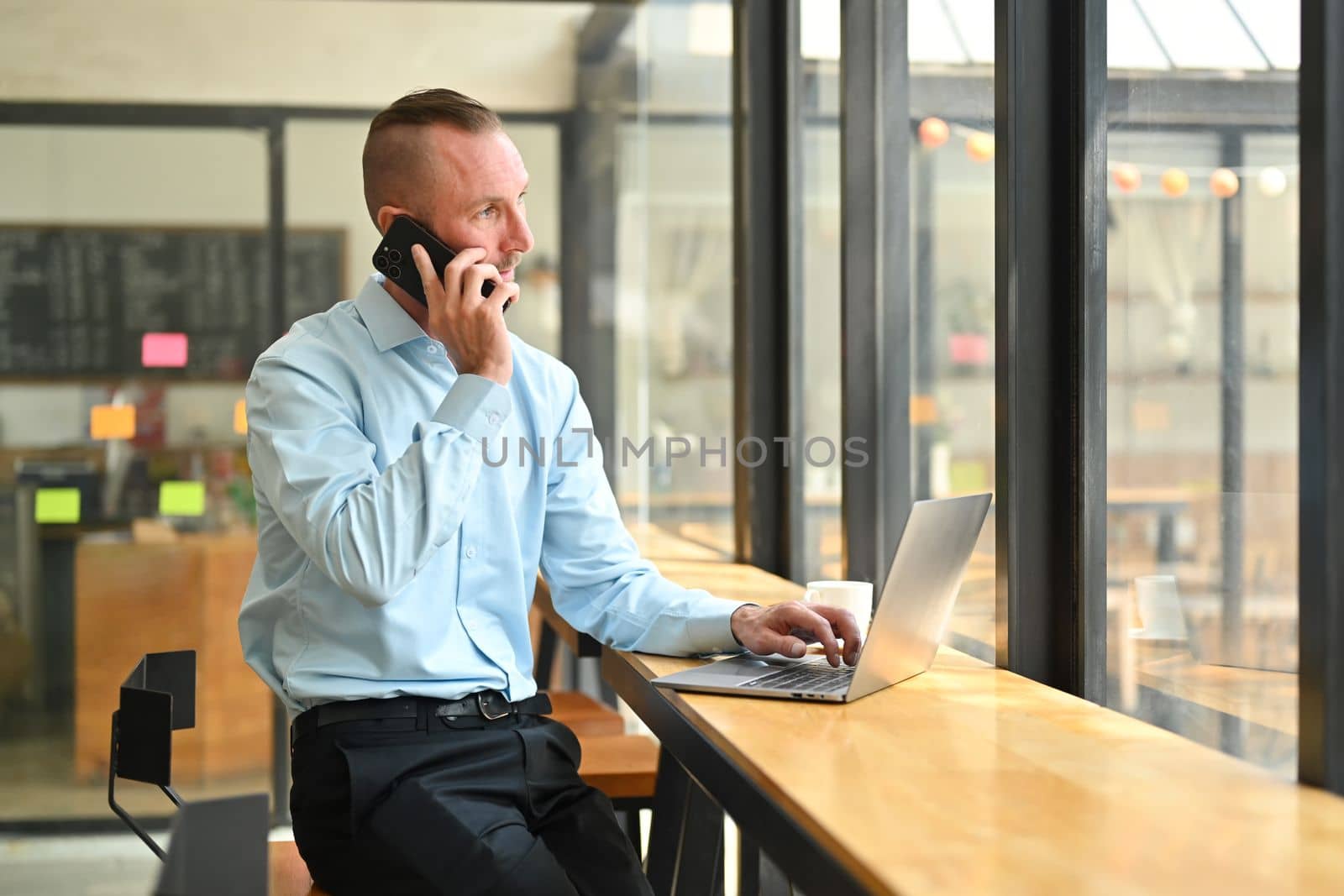 Portrait of businessman sitting at bright coffee shop talking on mobile phone and using laptop near glass window by prathanchorruangsak