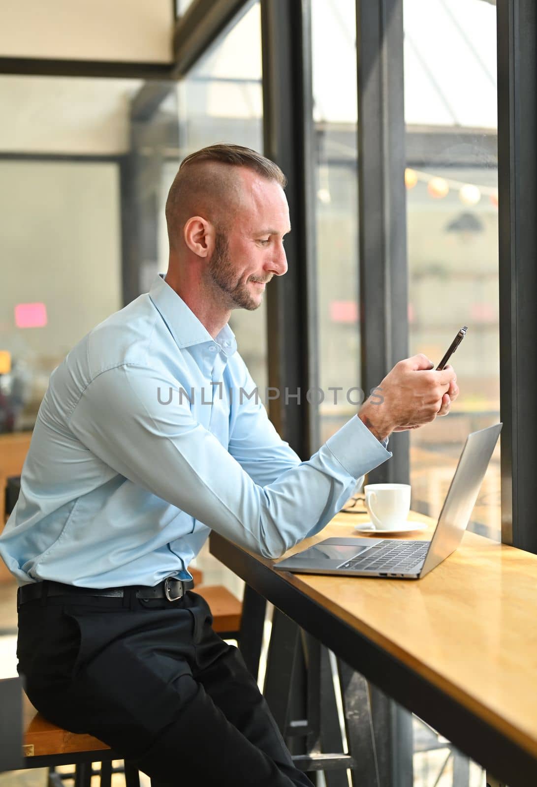 Portrait of millennial male entrepreneur sitting at wooden counter in modern cafe and using smart phone by prathanchorruangsak
