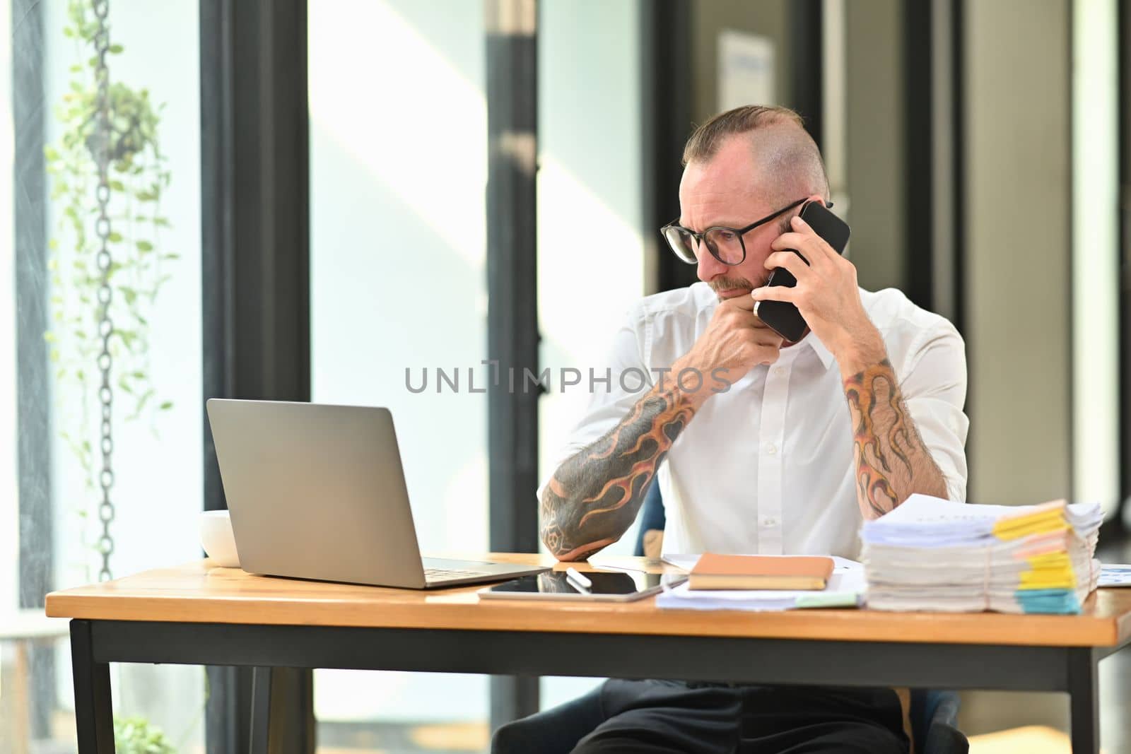 Handsome adult man employee in white shirt and glasses having phone conversation while sitting at desk in modern office by prathanchorruangsak