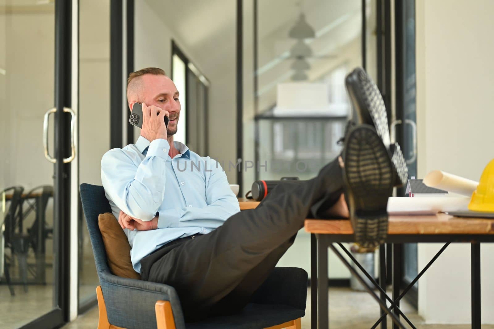 Happy caucasian businessman in shirt putting legs on table and talking on mobile phone in his personal office.
