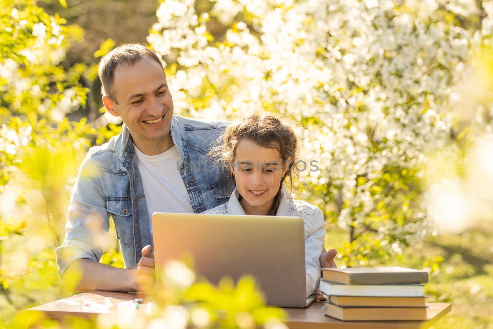 A girl and a young father are sitting at a laptop and studying in a flowered garden. Against the background of green grass and flowering trees. Remotely buy items in a store by Andelov13