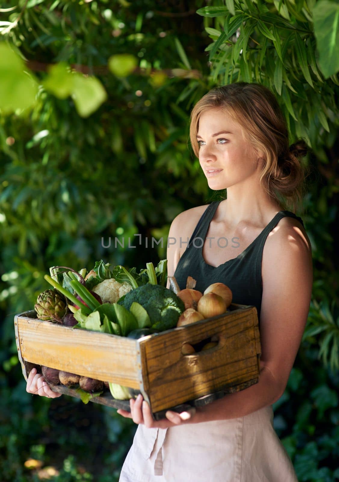 Shes down to earth. A young woman holding a crate of vegetables outdoors. by YuriArcurs