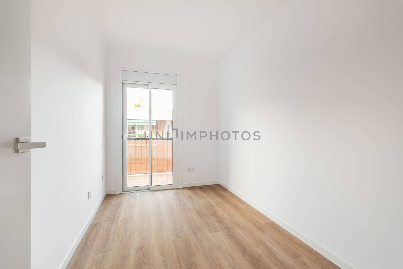 Bright room with white walls, light brown wooden parquet. The room has sliding glass doors in a metal frame with access to a balcony and a view of the neighboring house. Room for living
