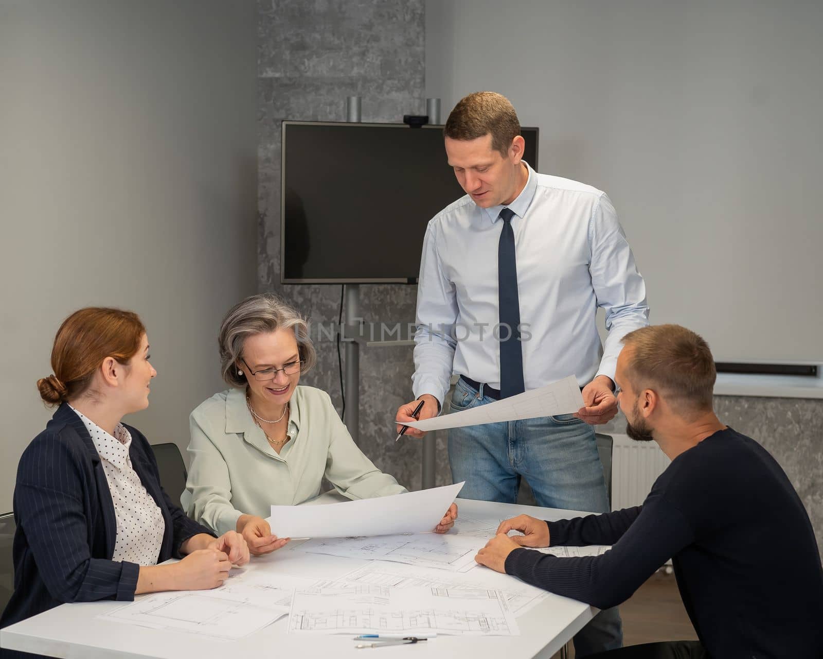 A caucasian man stands and holds a drawing, three colleagues sit at a table and listen to him