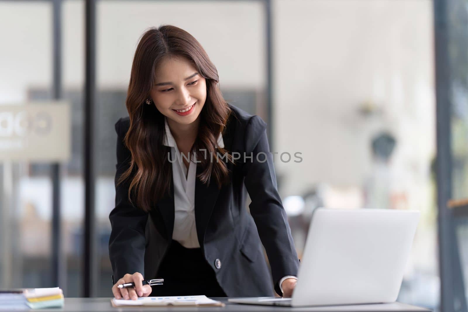 Young business woman standing in office writing work using laptop and take note by nateemee
