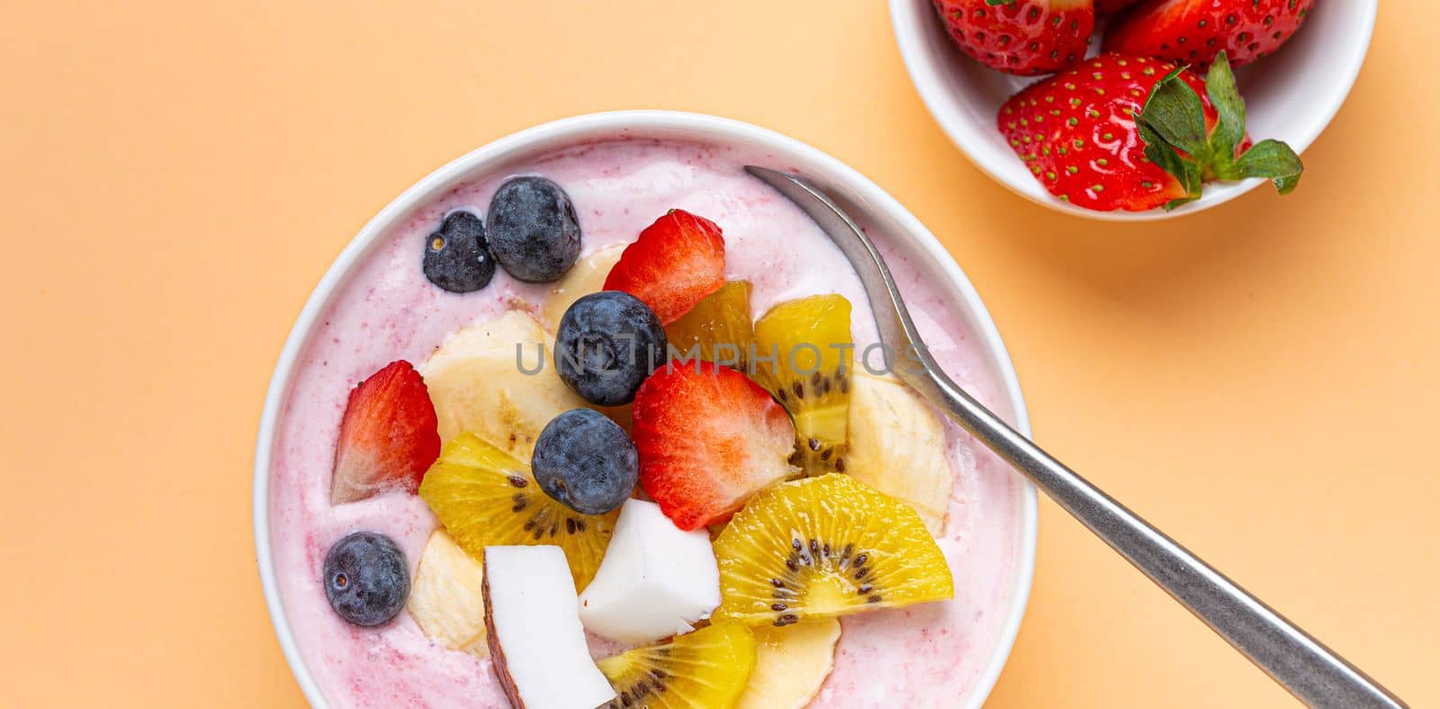 Healthy breakfast or dessert yogurt bowl with fresh banana, strawberry, blueberry, cocos, kiwi top view on minimal pastel paper background and spoon by its_al_dente