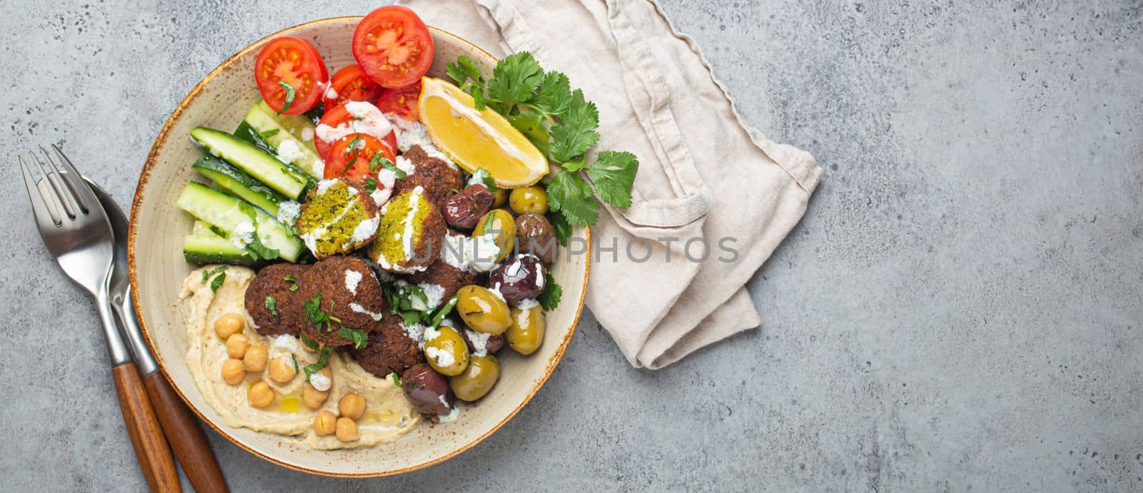 Falafel salad bowl with hummus, vegetables, olives, herbs and yogurt sauce. Vegan lunch plate top view on rustic stone background, healthy meal with falafel and veggies, copy space by its_al_dente