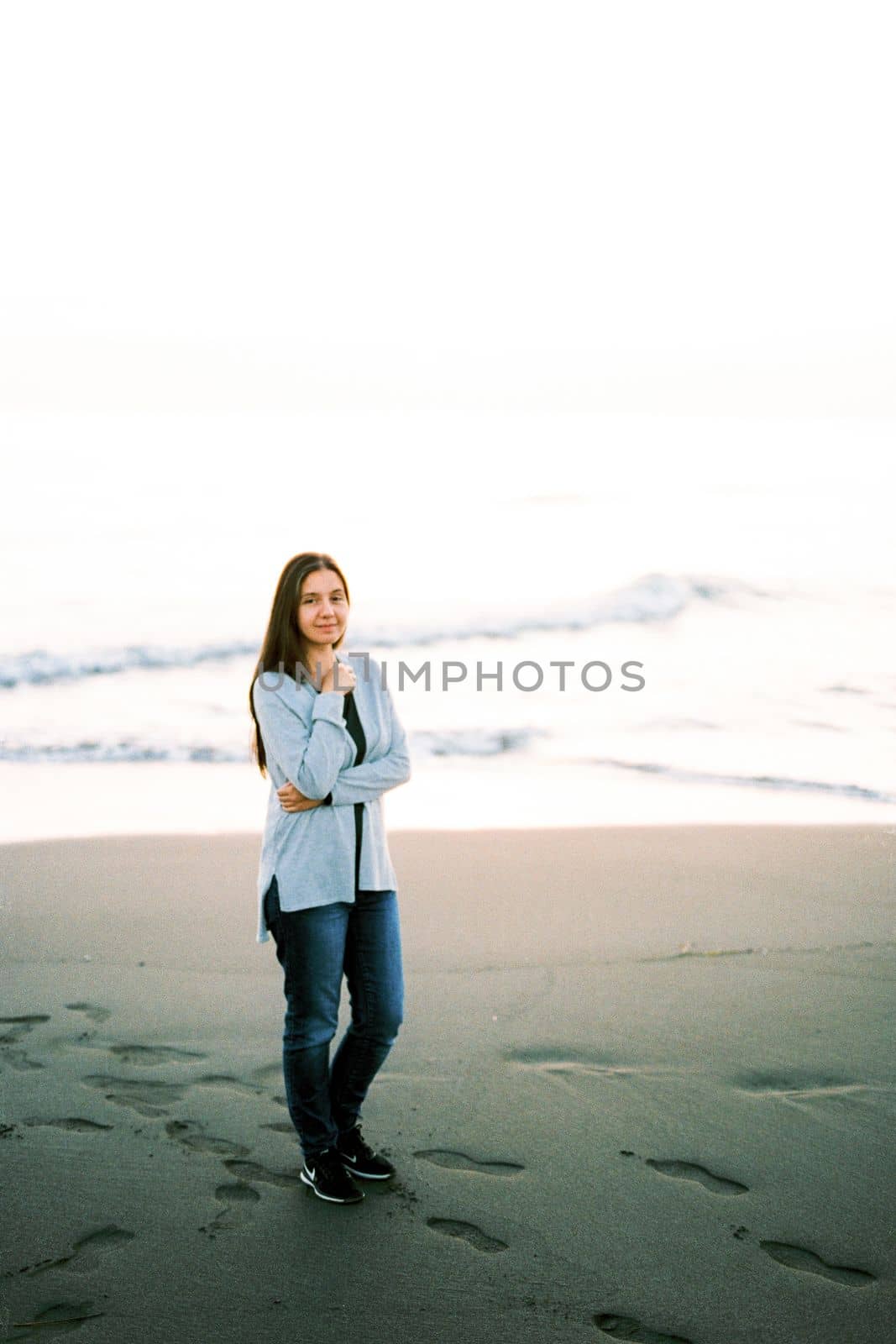 Young woman in a sweater and jeans stands on the beach with her arms crossed over her chest. High quality photo