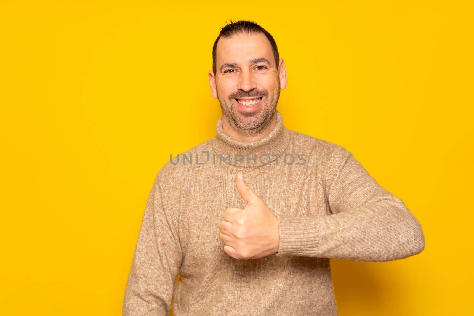 Bearded hispanic man wearing beige turtleneck posing with thumbs up in approval and self-confidence isolated over yellow background. Concept of success and positivism. by Barriolo82