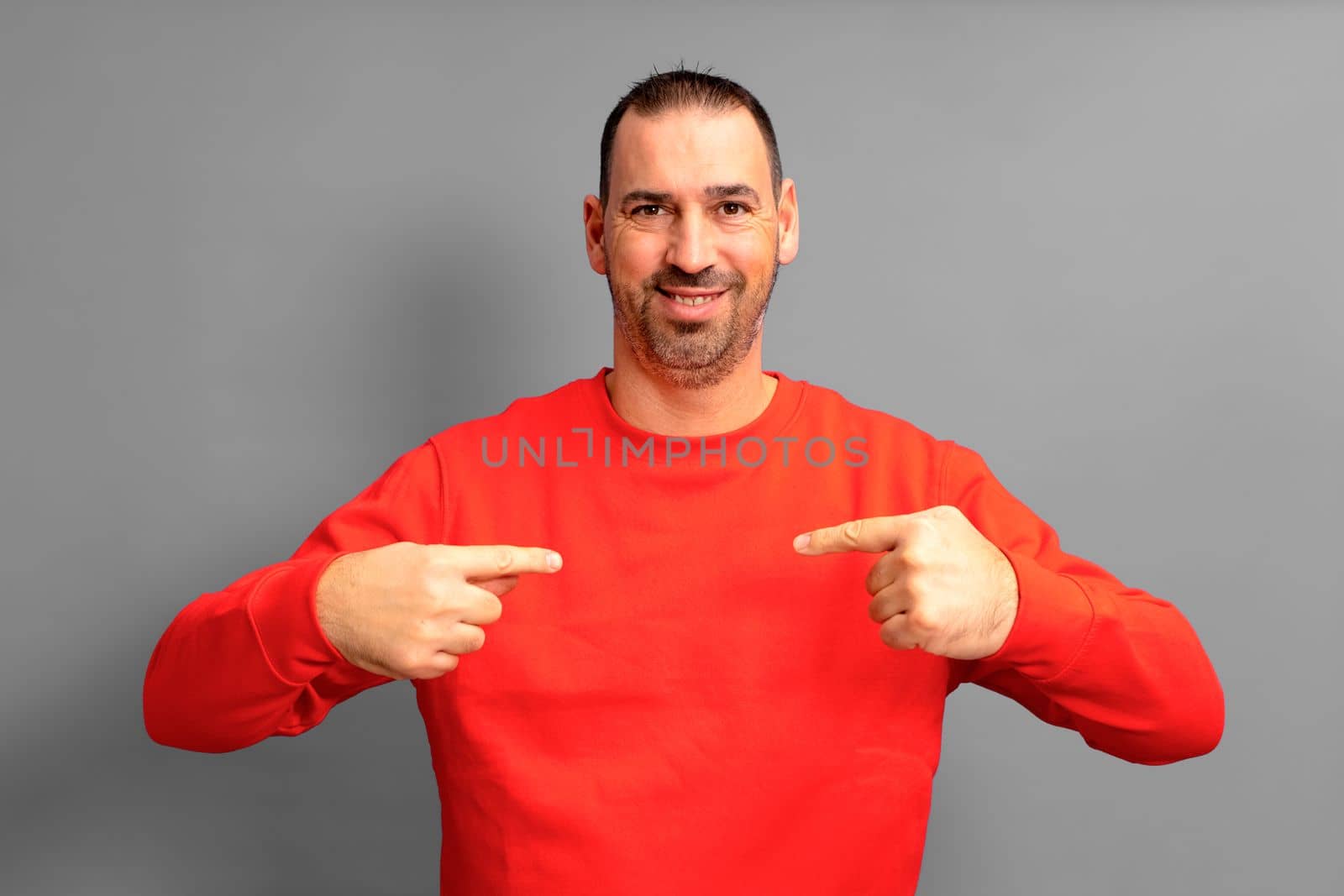 Bearded hispanic man wearing a red sweater pointing to himself proudly isolated on gray background. He is satisfied with his work, he feels effective and competitive