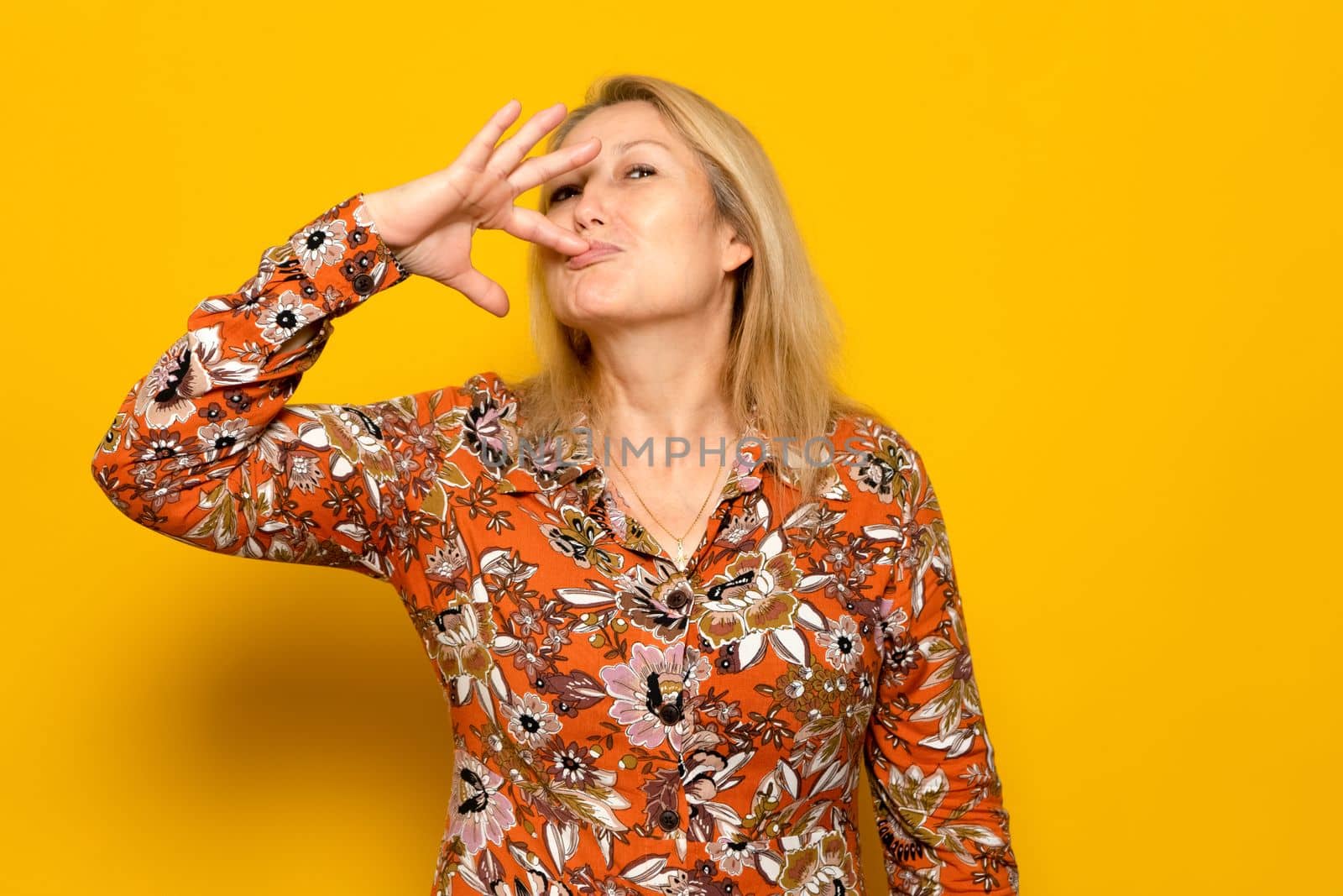 Caucasian blonde woman in a patterned dress fingering herself with pure pleasure isolated over yellow background, she has tasted delicious delicacies and is licking her lips with pleasure