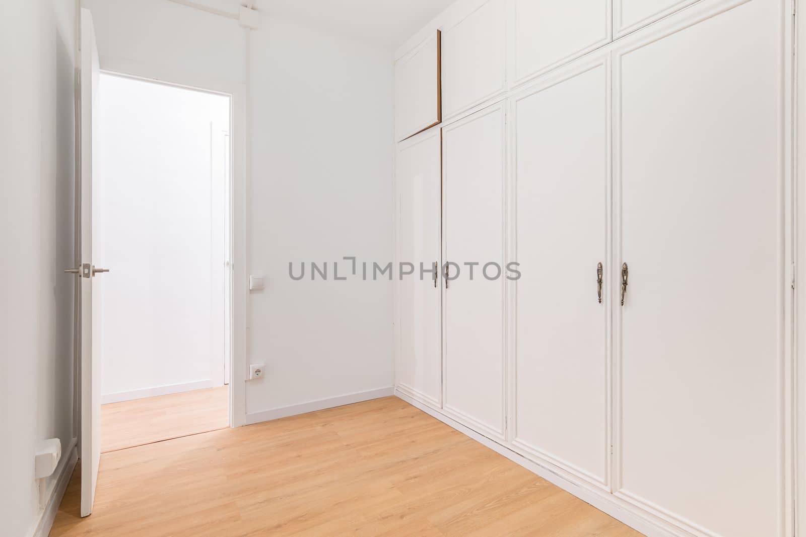 Built-in spacious wardrobes with white paneling and door open to another room with wooden laminate flooring. Concept of organizing storage and laconic interior.