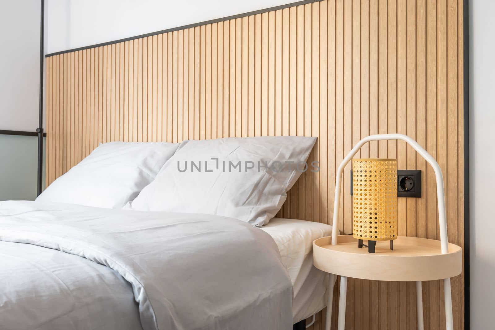 Closeup of a bedroom with a double bed against a white wall with designer wooden elements. On the bedside table is a night lamp with a yellow shade