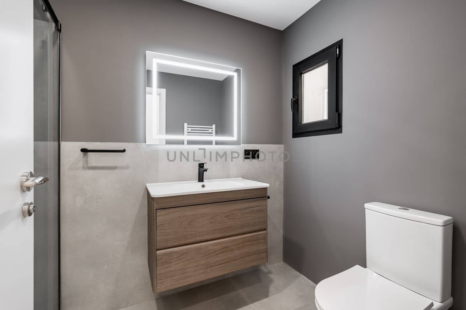Part of the bathroom in gray and white pastel colors with a matte white ceiling. Sink on a wooden countertop with drawers. On the tiled floor against the wall is a white toilet with a closed lid