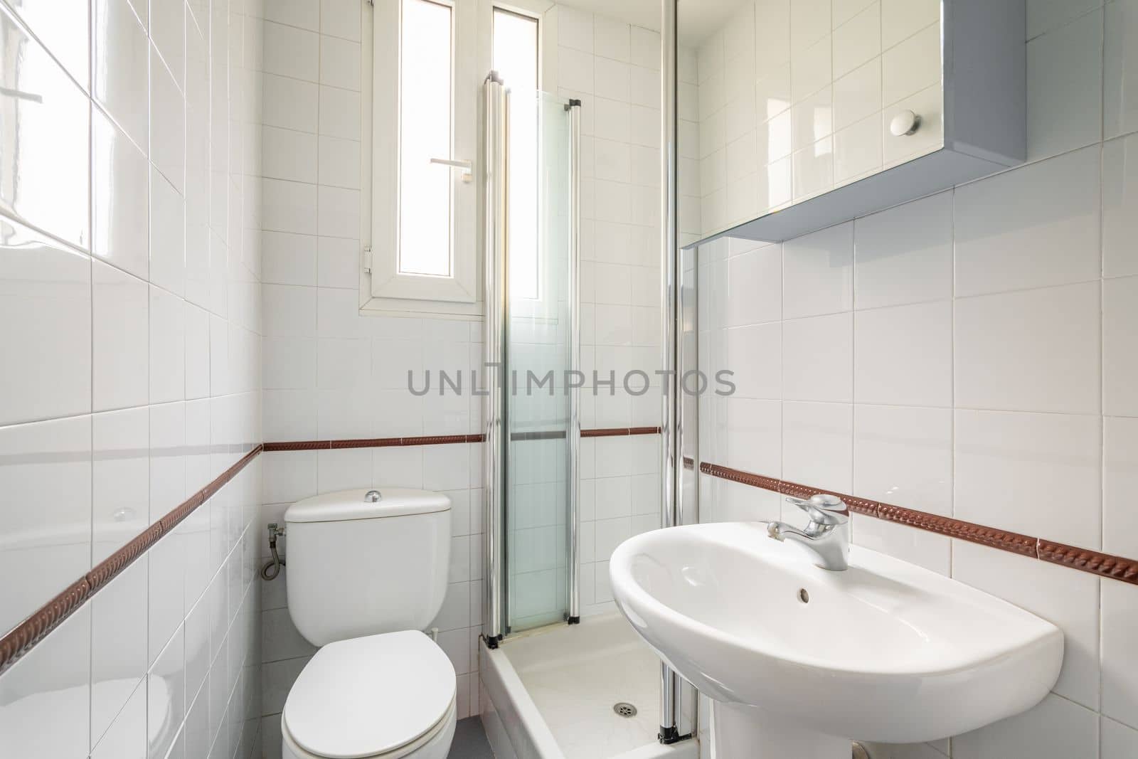 Luxury bathroom is lined with white ceramic tiles with shower toilet and sink with a narrow vertical windows for natural light. Concept of compact but comfortable bathrooms.