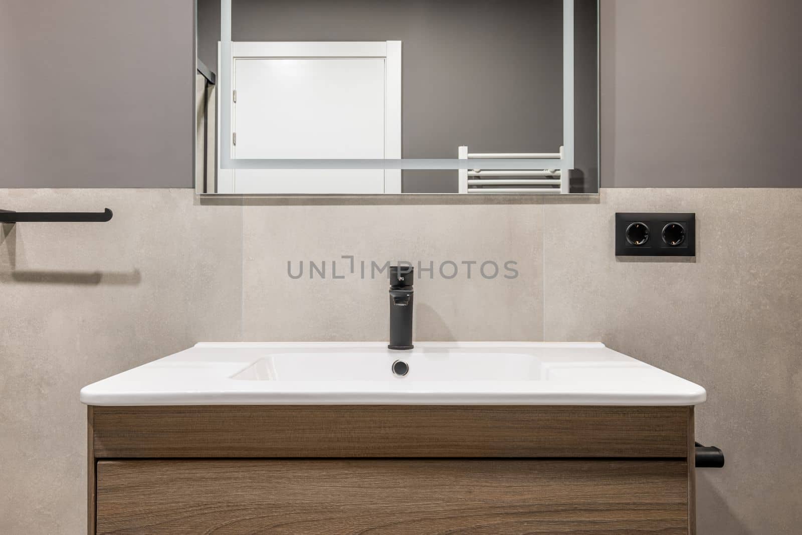 Square white ceramic sink on wooden table with drawers. On wall is mirror with white neon lighting. Mirror reflects white door and towel radiator. Walls are combination of marble tiles and gray paint
