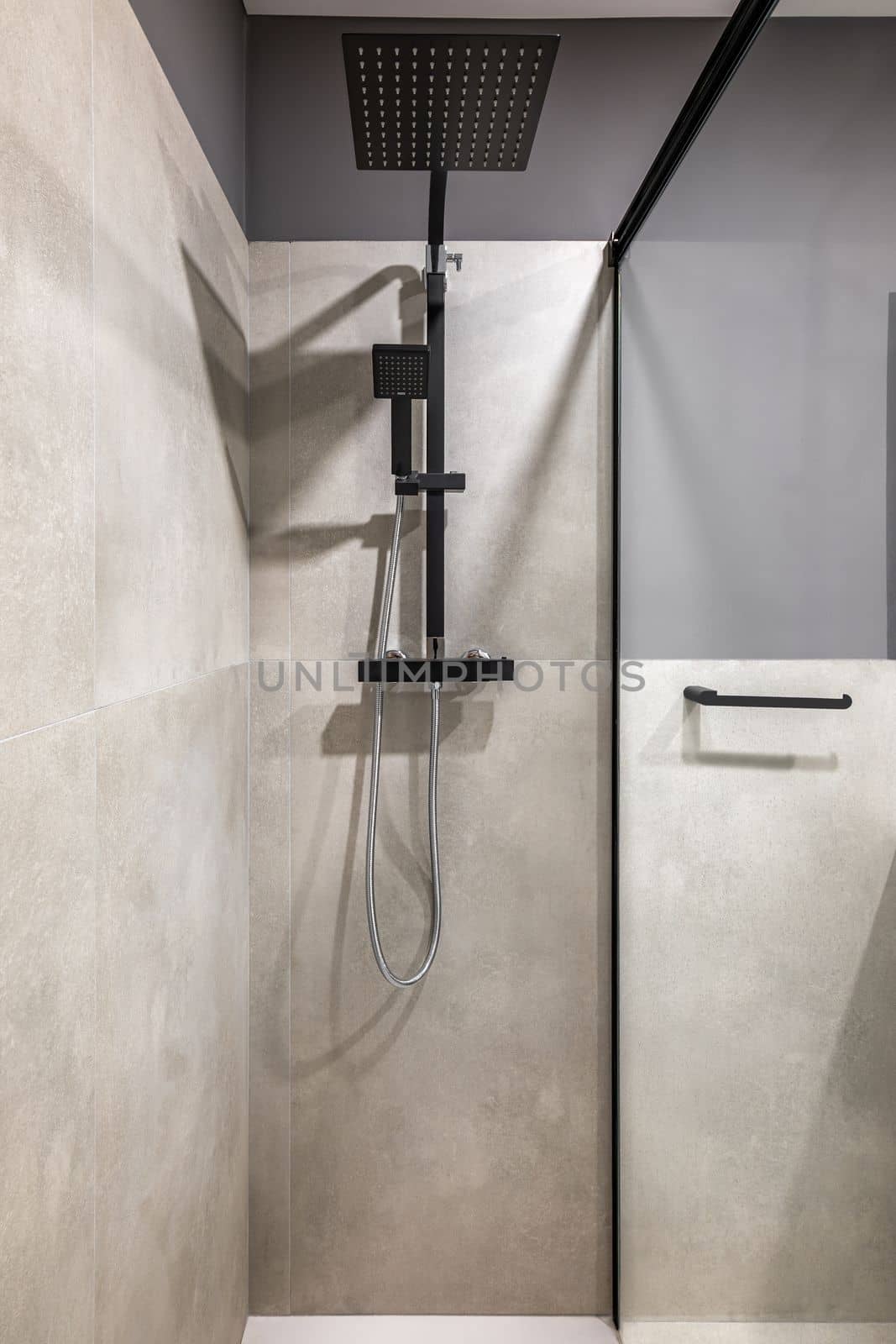Closeup of the shower area. Moisture resistant gray marble tile walls. On the wall is a black faucet with a large shower head. Splash guard made of durable clear glass with a black frame. by apavlin