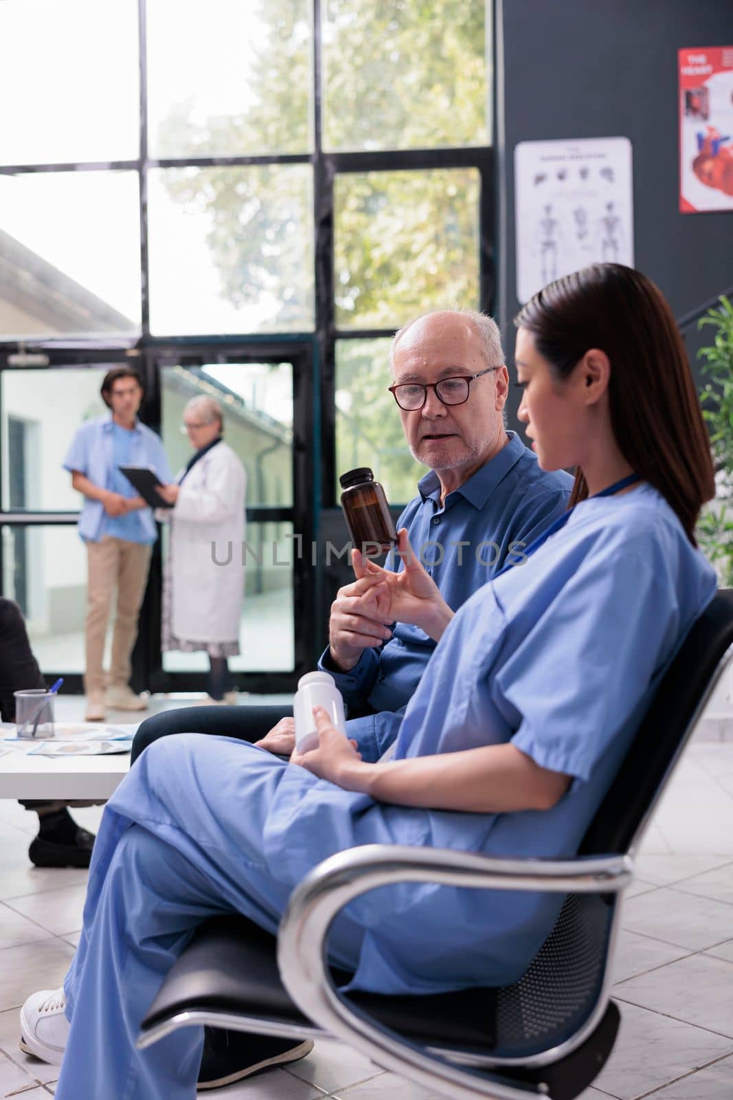 Medical assistant explaining medication treatment to elderly patient while showing pills bottle during consultation in hospital reception. Diverse people waiting in lobby before start examination