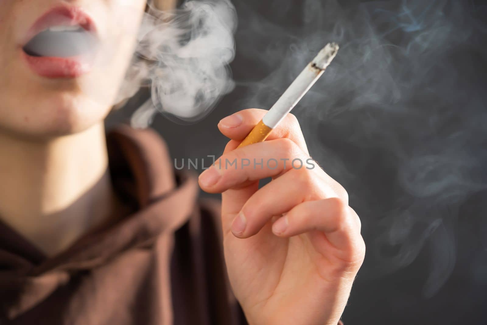 A young beauty girl is smoking, a woman is stressed and holds a cigarette in her hands, staying near the window of her apartment, close-up view.