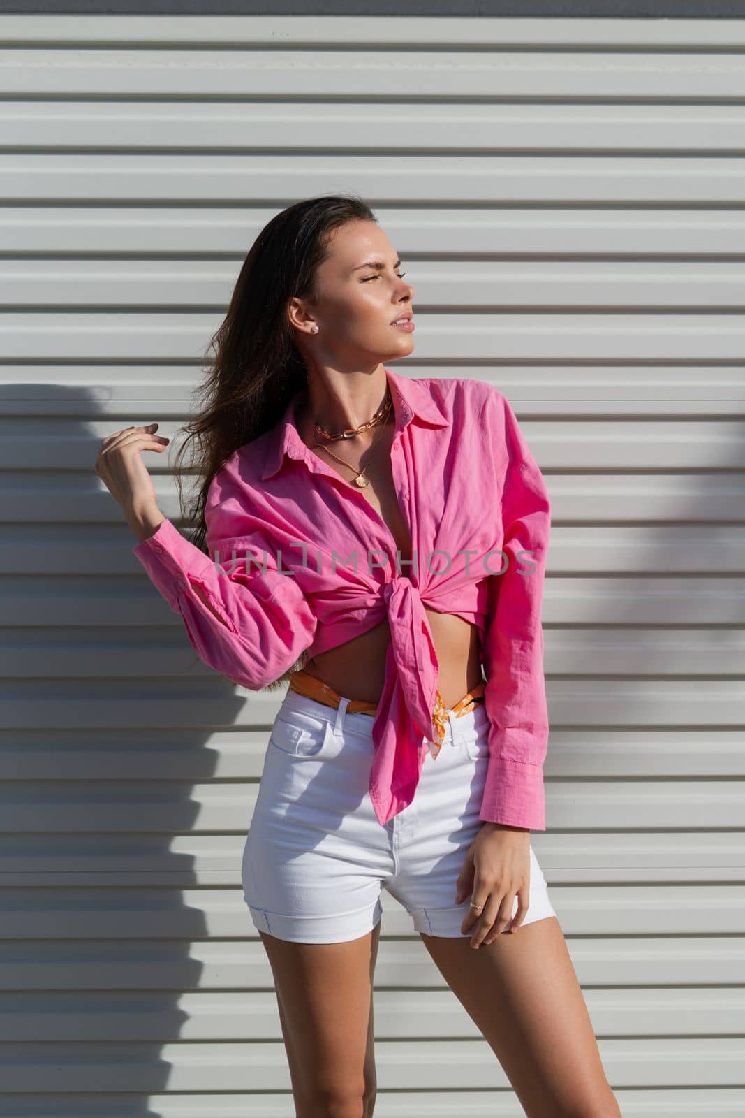 Young beautiful brunette woman in a pink shirt and white denim shorts against the background of a light garage door fence by kroshka_nastya