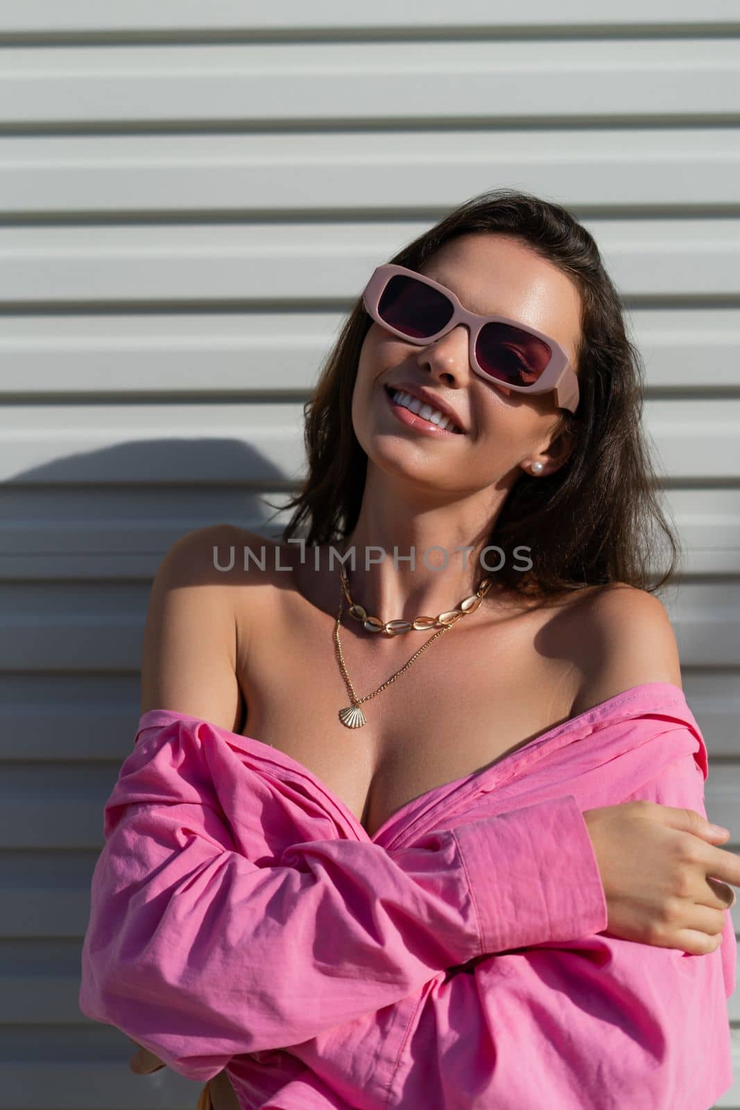 Young beautiful brunette in a pink shirt, neck jewelry, necklace, trendy sunglasses on the background of a light garage door fence