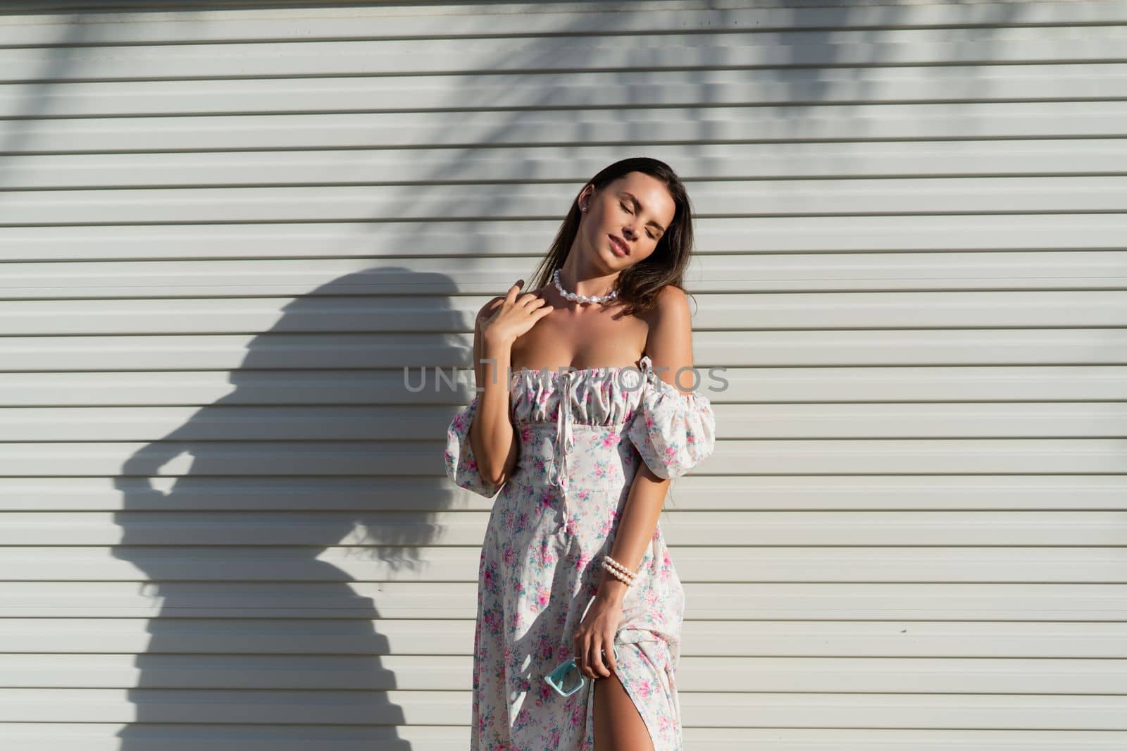 Young beautiful woman in a romantic dress with a floral print and a pearl necklace bracelet on the background of a light garage fence by kroshka_nastya