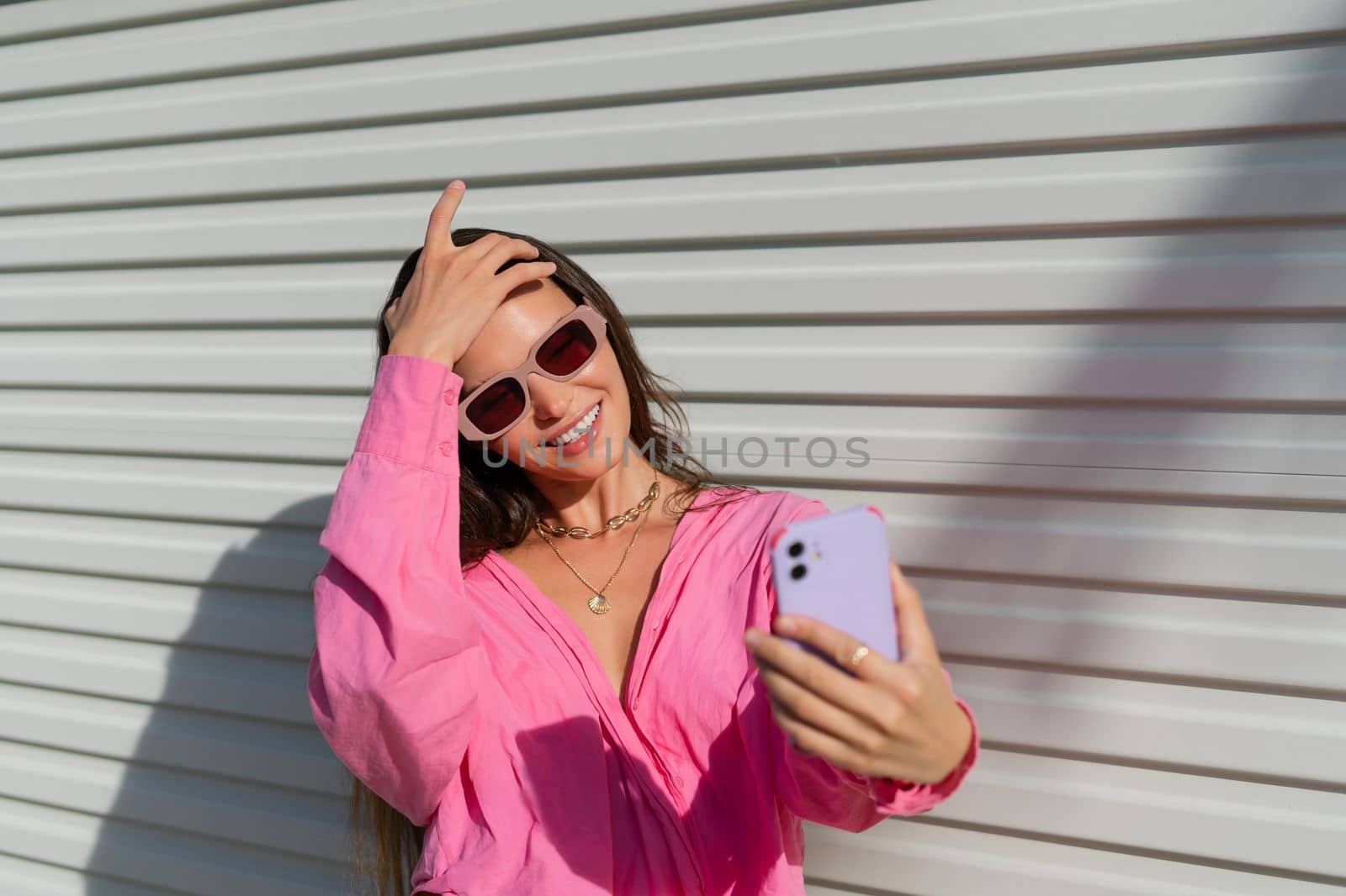 Young beautiful brunette in a pink shirt, neck jewelry, necklace, trendy sunglasses on the background of a light garage fence, takes a selfie on the phone, laughs, smiles by kroshka_nastya