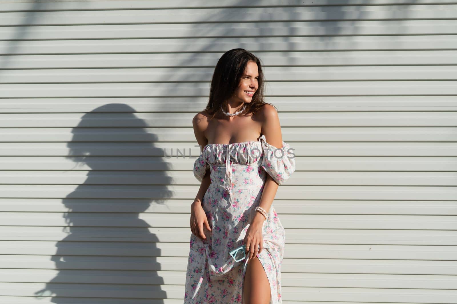 Young beautiful woman in a romantic dress with a floral print and a pearl necklace bracelet on the background of a light garage fence