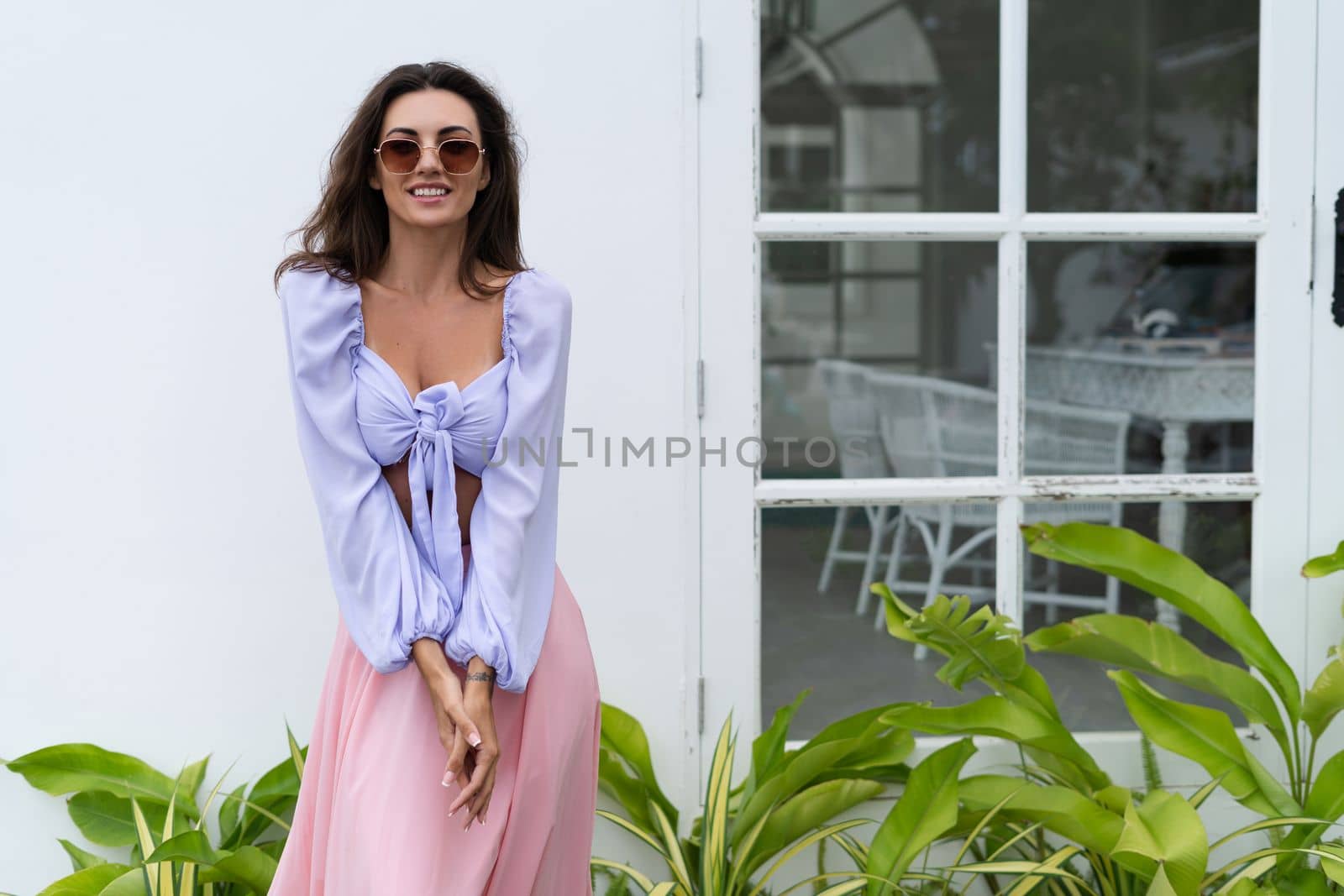 Pretty young stylish woman in trendy purple crop top long sleeve , pink skirt, isolated, white wall, tropical leaves natural day light..