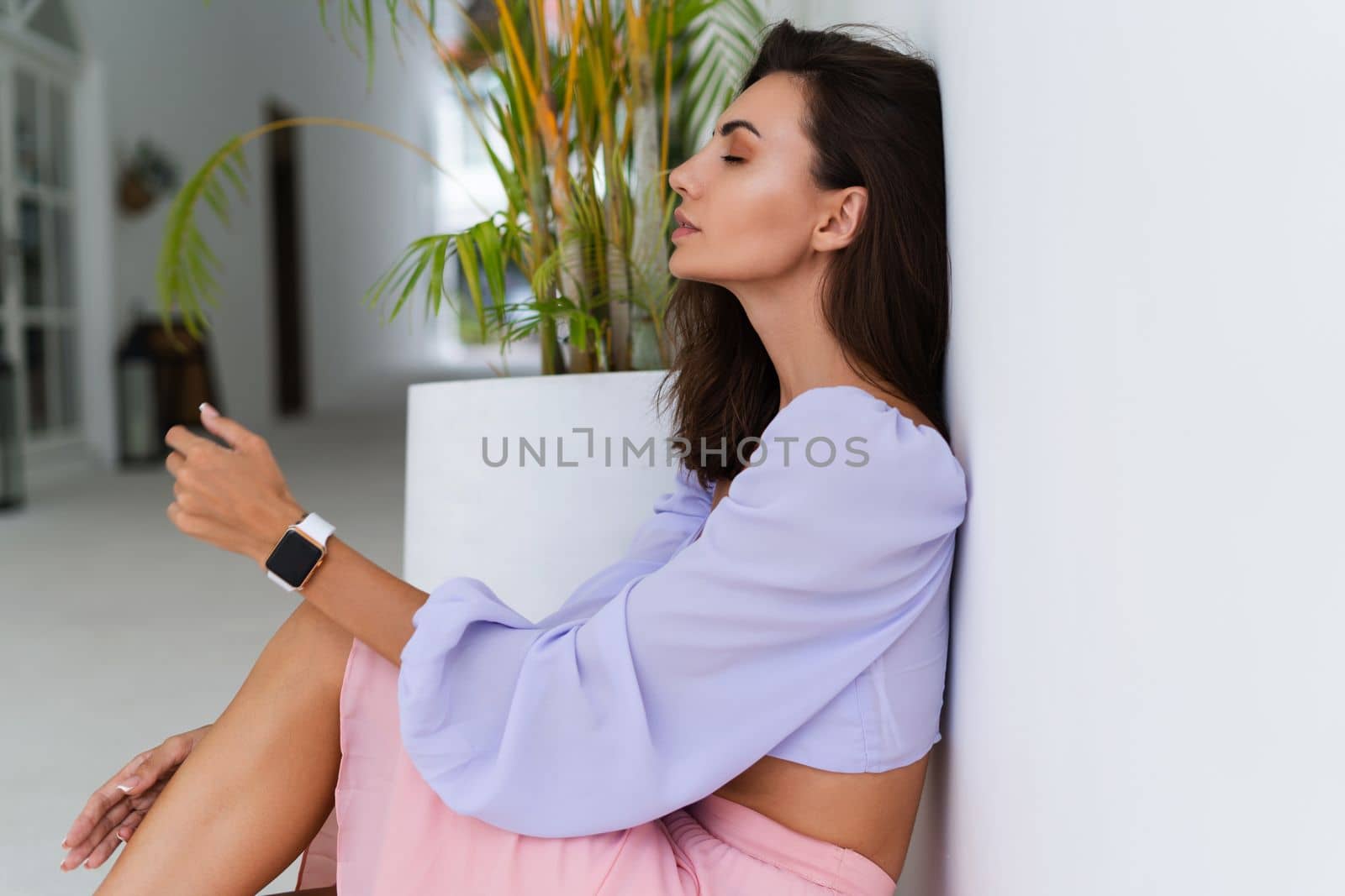 Stylish young woman with voluminous hair in a trendy long sleeve crop top and a pink skirt posing against a white wall and a tropical bush, wearing a smart watch