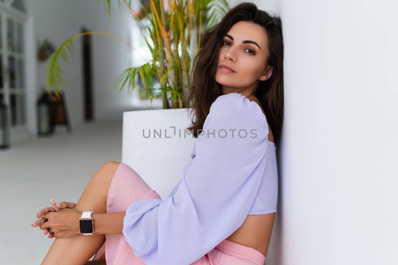 Stylish young woman with voluminous hair in a trendy long sleeve crop top and a pink skirt posing against a white wall and a tropical bush, wearing a smart watch by kroshka_nastya