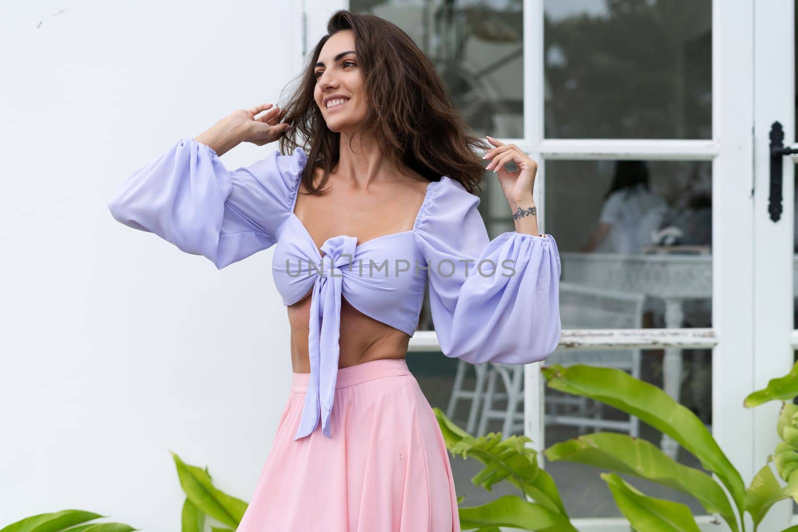 Pretty young stylish woman in trendy purple crop top long sleeve , pink skirt, isolated, white wall, tropical leaves natural day light..