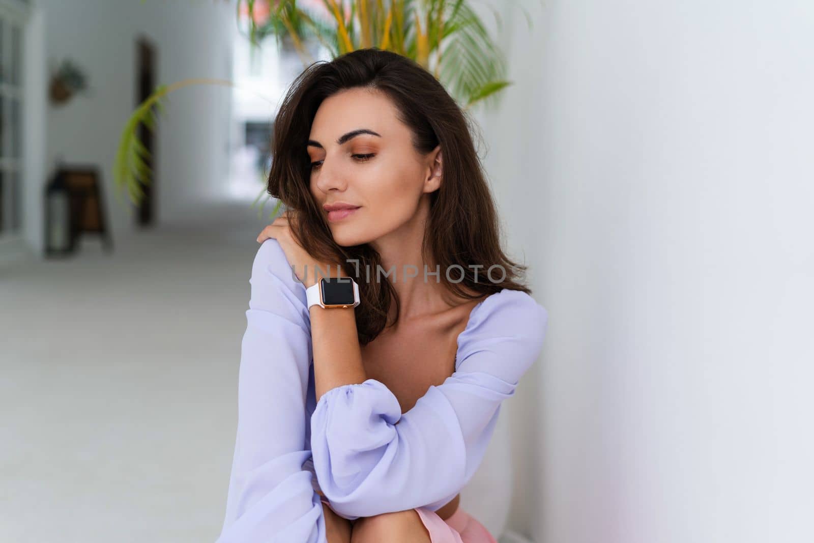 Stylish young woman with voluminous hair in a trendy long sleeve crop top and a pink skirt posing against a white wall and a tropical bush, wearing a smart watch