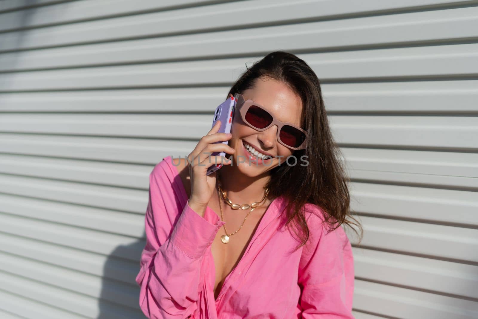 Young beautiful brunette woman in a pink shirt, neck jewelry, necklace, trendy sunglasses on the background of a light garage fence, calls the phone, laughs, smiles