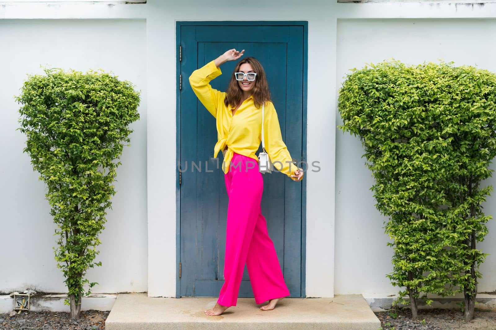 Stylish fit fashion women in bright pink wide leg pants and yellow shirt holding bag trendy mint sunglasses posing at luxury tropical villa by pool outdoor natural day light