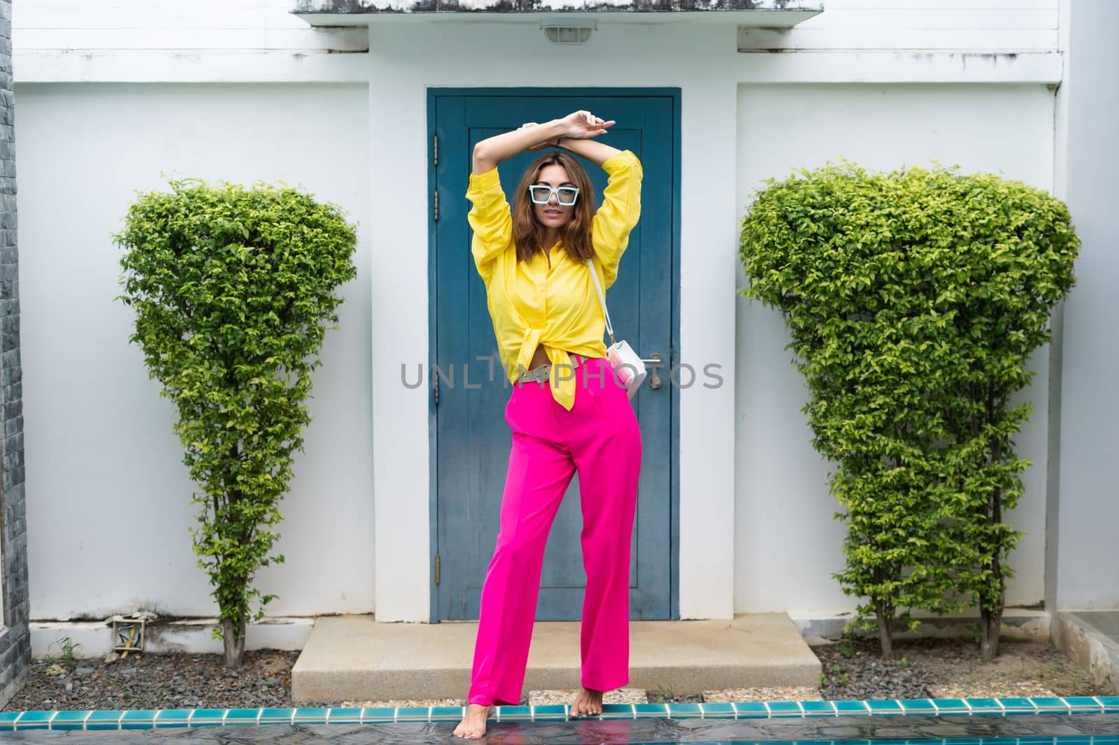 Stylish fit fashion women in bright pink wide leg pants and yellow shirt holding bag trendy mint sunglasses posing at luxury tropical villa by pool outdoor natural day light